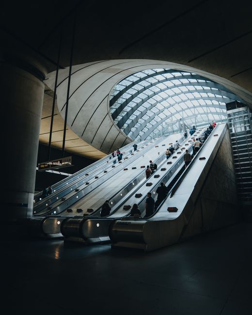 Inside a modern transportation hub, a large, curved glass and steel structure features an expansive, naturally lit roof with a geometric pattern. A wide, multi-lane escalator with metallic handrails a