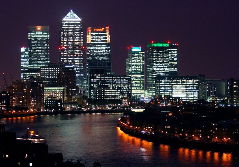 A low-angle view of three modern office buildings in Canary Wharf, featuring glass facades and steel framing, with the middle building being a tall, white rectangular skyscraper with a grid pattern of