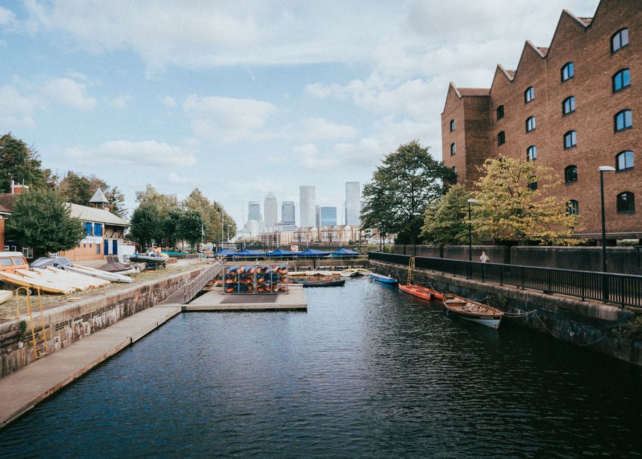 A wide view of the Canary Wharf skyline under a clear blue sky, featuring modern high-rise office buildings with glass facades and steel structures, situated behind a row of low-rise residential or co