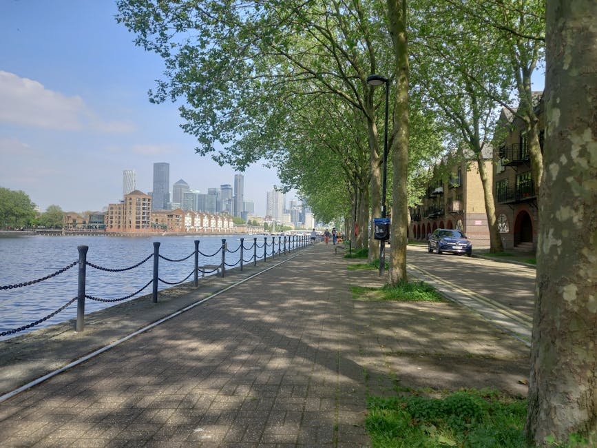 A wide pavement running alongside the east London River Thames, with a row of mature green trees casting dappled shade on the pathway. To the left, a black metal chain-link fence separates the pedestrian walkway from the water, where several small boats are moored along the riverbank. In the background, modern high-rise office buildings of Canary Wharf rise against a partly cloudy sky, indicating the urban skyline. On the right side of the image, there are residential buildings with brick facades, balconies, and parked cars along a narrow street. A black car is visible parked adjacent to the sidewalk, and there is a blue mailbox attached to a lamppost. The scene is during daylight, with natural lighting and shadows from the trees. This setting depicts an urban riverside environment suitable for a house or home relocation process, with the presence of commercial and residential infrastructure supporting moving logistics. Man and Van Canary Wharf provides removals services in this area.